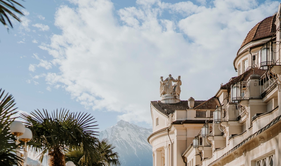 L'edificio dell'hotel con torre e balconi si trova davanti a uno sfondo di montagne sotto un cielo azzurro.