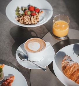 Ein Cappuccino mit Herzmuster, Croissant, Orangensaft und Schale mit Obst und Müsli auf dem Frühstückstisch.