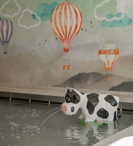 Ein kleiner Kuhbrunnen sprudelt Wasser in ein Becken vor einer Wand mit Heißluftballons.