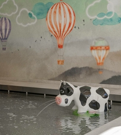 Ein kleiner Kuhbrunnen sprudelt Wasser in ein Becken vor einer Wand mit Heißluftballons.