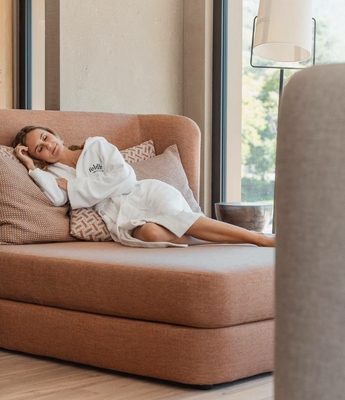 Frau im Ruhebereich des Wellnesshotels Eine Frau im Bademantel liegt entspannt auf einer Liege im Ruhebereich neben einem Fenster.