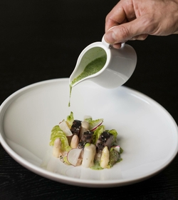 Eine Hand gießt grüne Sauce über ein kunstvoll angerichtetes Gourmetgericht in einer weißen Schale.