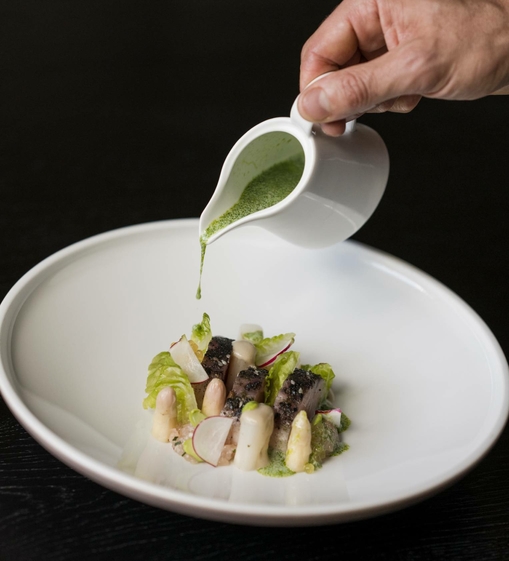 Eine Hand gießt grüne Sauce über ein kunstvoll angerichtetes Gourmetgericht in einer weißen Schale.