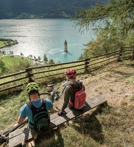 Two mountain bikers sit on a bench above the lake and look out over the water toward a tower in the lake.