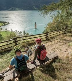 Two mountain bikers sit on a bench above the lake and look out over the water toward a tower in the lake.