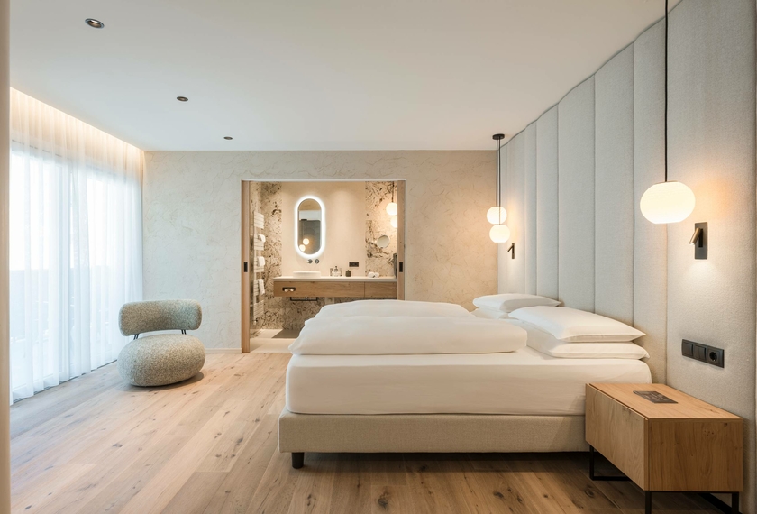 Ein helles Doppelzimmer mit zwei Betten, Holzboden und einem offenen Blick ins Badezimmer.