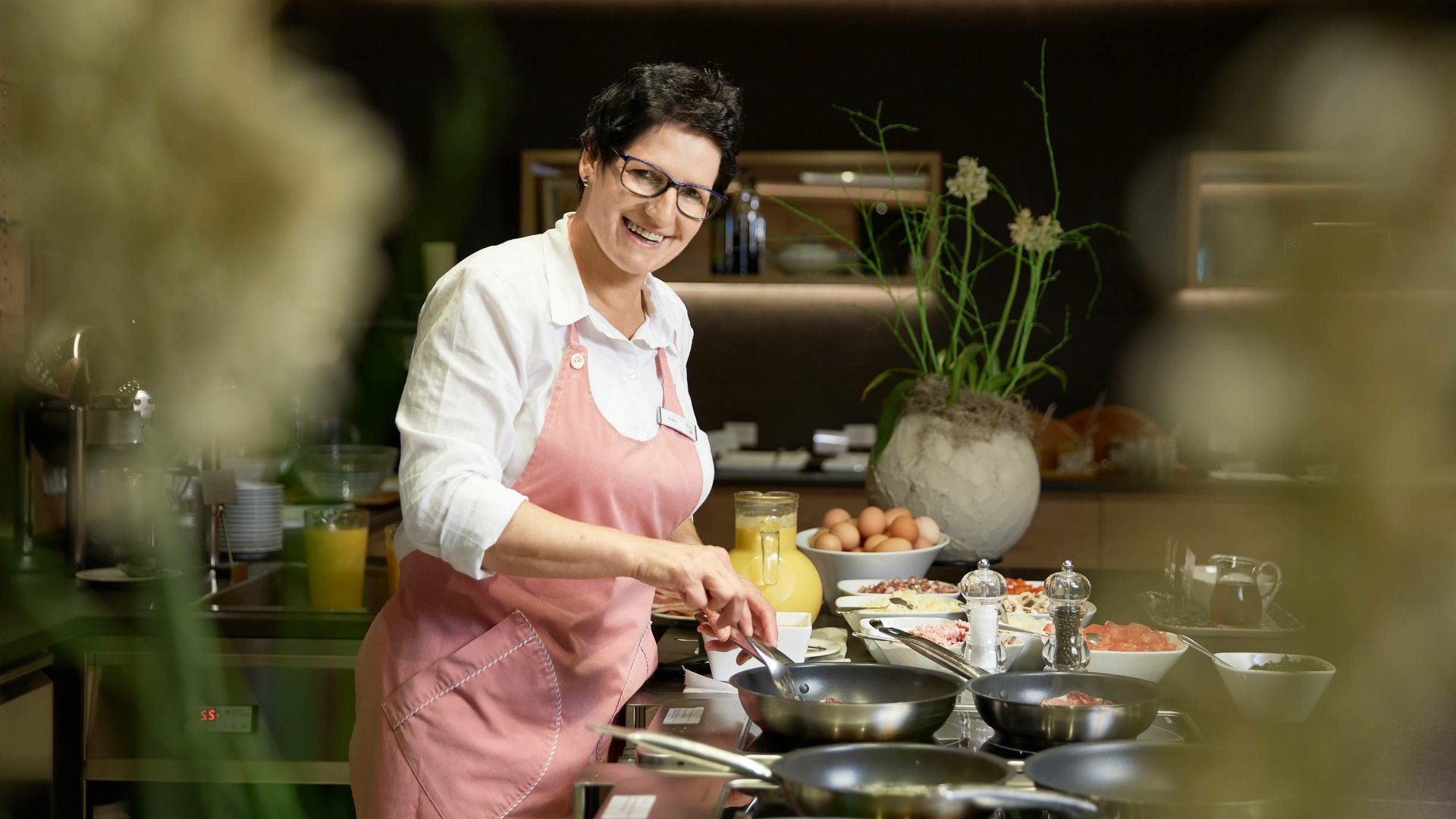 A female chef in a pink apron prepares dishes at the stove in the hotel restaurant kitchen.
