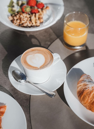A breakfast table shows a cappuccino, a croissant, orange juice and a bowl of cereal with fruit.