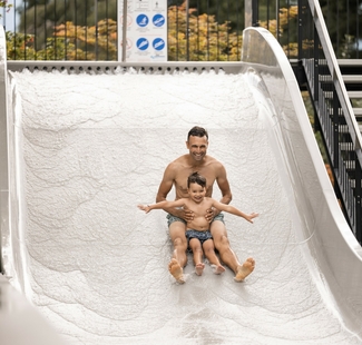 Un uomo e un bambino scendono su uno scivolo acquatico chiaro nell’area Family Spa, con il bambino a braccia aperte.