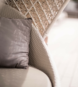 A rattan hanging chair with a grey cushion stands on a bright wooden terrace.