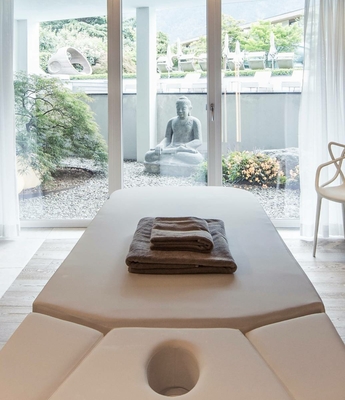 Massagebett im Spa des Feldhof in Naturns Ein Massagebett mit gefalteten Handtüchern steht vor großen Glasfenstern im Spa.