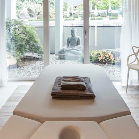 Tavolo per massaggi nella spa del Feldhof a Naturno Un lettino da massaggio con asciugamani piegati si trova davanti a grandi vetrate nella spa.