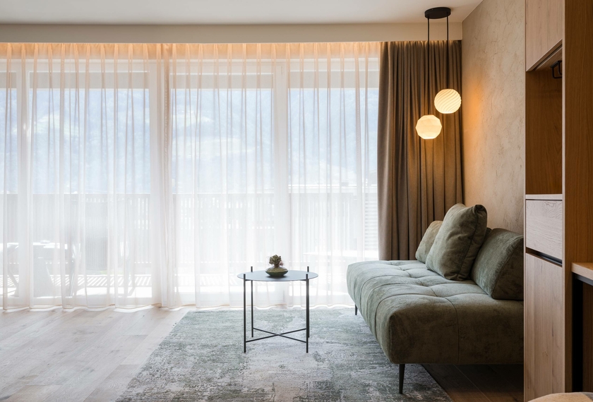 Suite Harmonie mit Balkon und Sitzbereich Ein heller Wohnbereich der Suite Harmonie mit Sofa, kleinem Tisch und bodentiefen Vorhängen vor dem Balkonfenster.