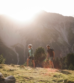 Zwei Wanderer gehen über eine Almwiese vor einer steilen Bergkulisse im warmen Abendlicht.