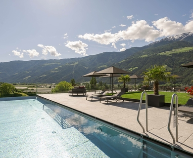 Infinity-Pool mit Bergblick im Hotel Naturns Ein Außenpool mit Liegen und Sonnenschirmen eröffnet den Blick auf die Berge bei klarem Himmel.