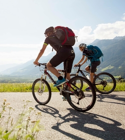 Two mountain bikers ride along a road overlooking the valley and mountains.