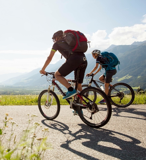 Two mountain bikers ride along a road overlooking the valley and mountains.