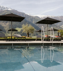 Una piscina a sfioro con lettini e ombrelloni davanti a un panorama montano e cime innevate.