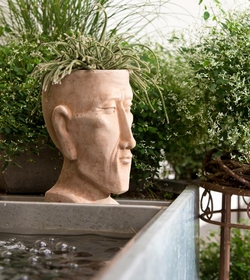 Eine dekorative Skulptur steht neben einem Becken mit Wasser und ist von grünen Pflanzen umgeben.