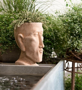 Eine dekorative Skulptur steht neben einem Becken mit Wasser und ist von grünen Pflanzen umgeben.