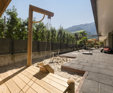 Spielplatz mit Sandbereich im Hotelgarten Ein kleiner Spielbereich mit Sandkasten, Holzpumpe und gepflasterter Terrasse liegt neben dem Gebäude mit Bergblick.