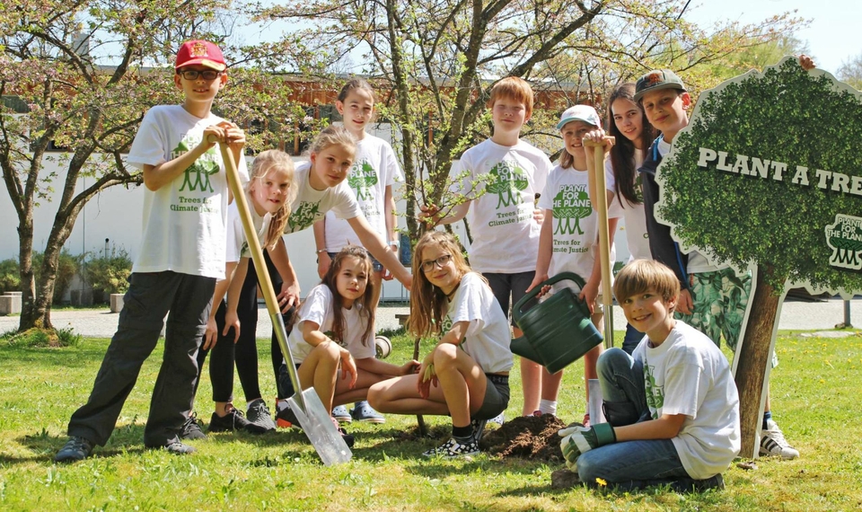 Several children stand and kneel in the garden holding shovels, seedlings and a watering can beside a tree-planting sign.