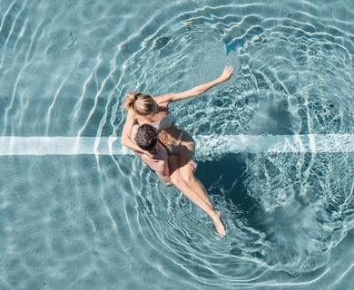 Pool im Feldhof DolceVita Resort Zwei Personen sitzen im Pool und umarmen sich, während sich Wasserringe um sie ausbreiten.
