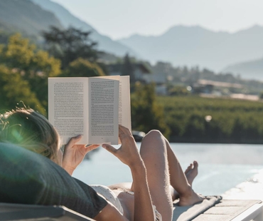 A person is reading on a lounger by the pool with mountains in the background.