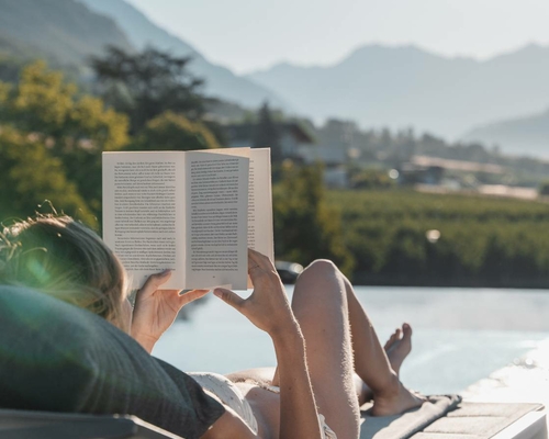 A person is reading on a lounger by the pool with mountains in the background.