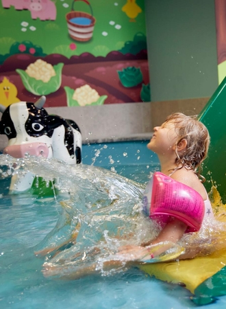 Ein Kind mit Schwimmflügeln sitzt an einer Wasserrutsche im Family Spa, während Wasser ins Becken spritzt.
