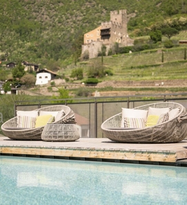 Due poltrone lounge con cuscini sono accanto alla piscina, con paesaggio montano e castello sullo sfondo.