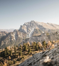 Wanderer auf einem alpinen Hang mit Blick auf ein felsiges Bergmassiv und Wald im Meraner Land.