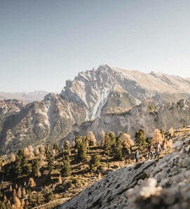 Wanderer auf einem alpinen Hang mit Blick auf ein felsiges Bergmassiv und Wald im Meraner Land.