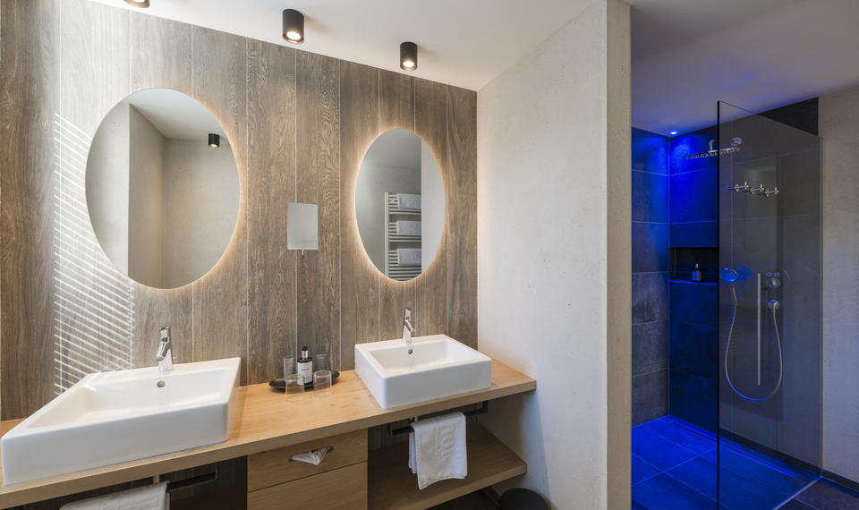 Suite-Badezimmer mit Doppelwaschbecken und Dusche Ein modernes Badezimmer mit Doppelwaschbecken, ovalen Spiegeln und einer blau beleuchteten Glasdusche.