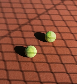 Two tennis balls lie on a tennis court with the shadow of a net across the surface.