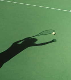 The shadow of a tennis player reaches toward the ball on the green court.