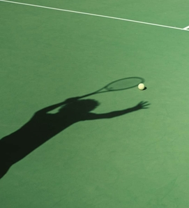 The shadow of a tennis player reaches toward the ball on the green court.