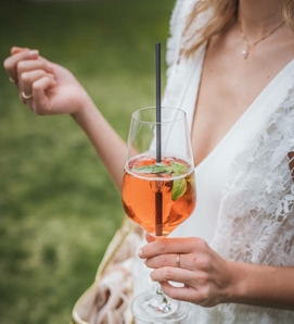 Eine Frau hält ein Glas mit orangefarbenem Cocktail und Strohhalm, im Hintergrund ist eine grüne Wiese zu sehen.