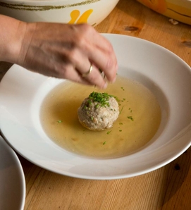 Eine Hand bestreut einen Knödel in klarer Brühe mit Kräutern in einem tiefen Teller.