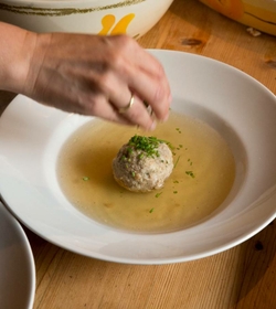 Eine Hand bestreut einen Knödel in klarer Brühe mit Kräutern in einem tiefen Teller.