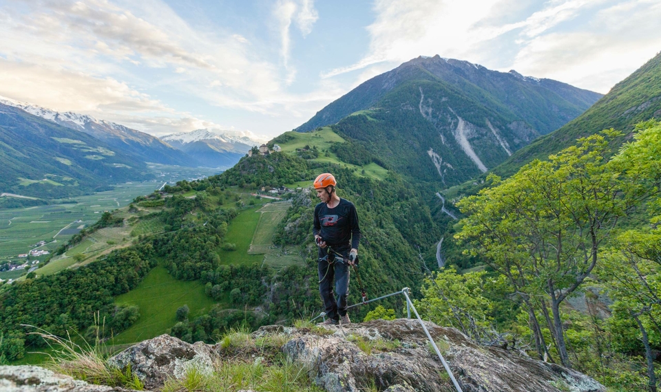 Una persona con casco e imbrago da arrampicata sta su una roccia davanti a un paesaggio montano verde.