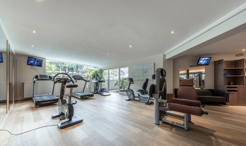 Ein heller Fitnessraum mit mehreren Trainingsgeräten, Spiegelwand und großen Fenstern.