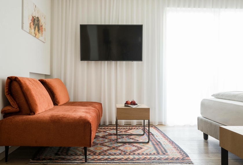 Ein modernes Hotelzimmer mit orangefarbenem Sofa, TV, kleinem Tisch und hellem Tageslicht am Fenster.