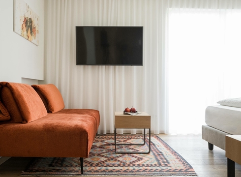 Ein modernes Hotelzimmer mit orangefarbenem Sofa, TV, kleinem Tisch und hellem Tageslicht am Fenster.