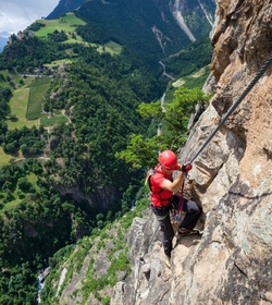 Eine Person klettert gesichert an einer steilen Felswand mit Blick ins grüne Tal in Südtirol.