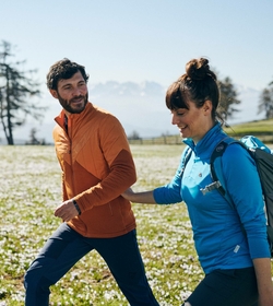 Ein Paar wandert bei Sonnenschein über eine blühende Wiese vor alpiner Bergkulisse.