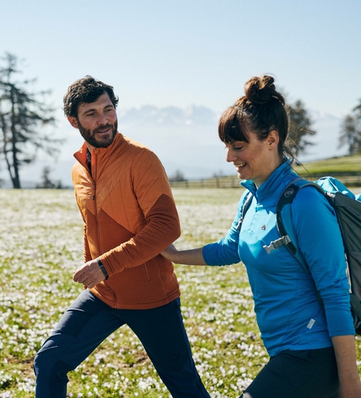 Ein Paar wandert bei Sonnenschein über eine blühende Wiese vor alpiner Bergkulisse.