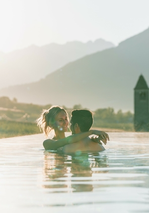 Una coppia si abbraccia nella piscina a sfioro con le montagne sullo sfondo e la luce della sera.