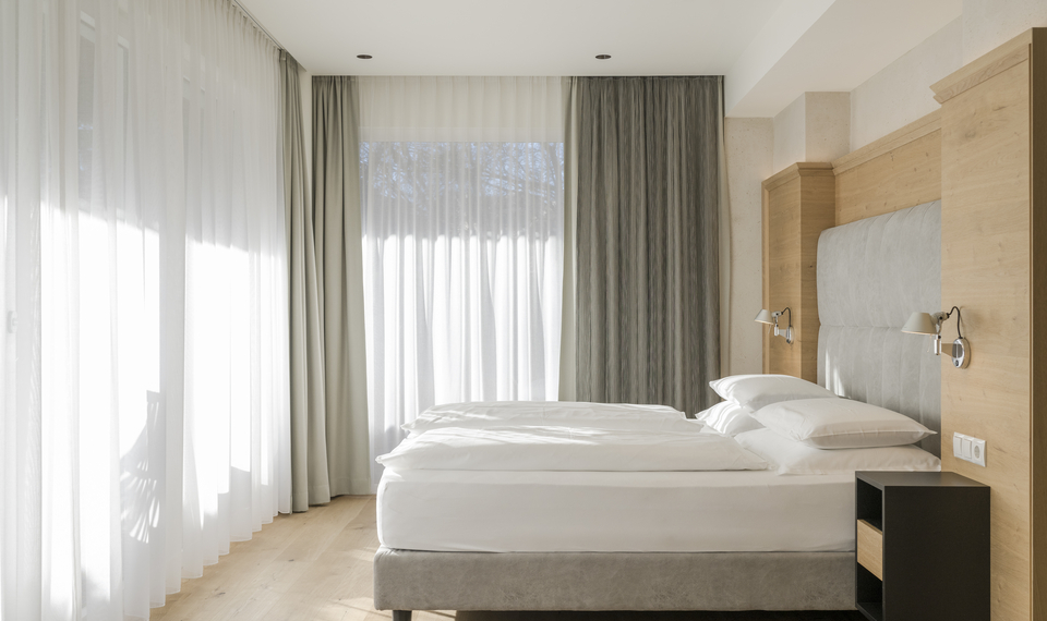 Suite mit Balkon im Feldhof DolceVita Resort Ein helles Schlafzimmer einer Suite mit großem Bett, Holzdetails und bodentiefen Vorhängen.
