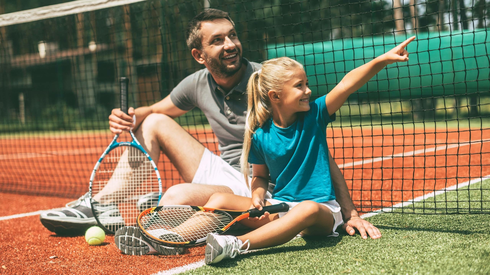 A man and a girl sit on a tennis court with rackets and smile while pointing to the right.